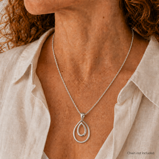 A close-up of a woman wearing a silver double teardrop pendant necklace with a silver chain, styled with a light beige linen shirt. The text 'Chain not included' is visible in the bottom right corner.